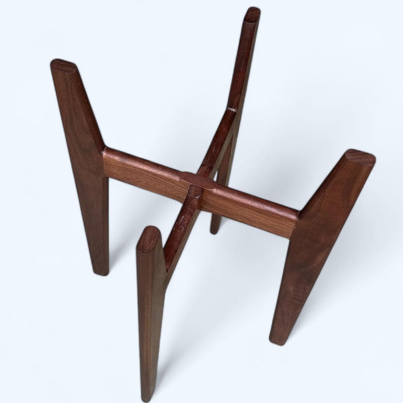 May include: A dark brown wooden plant stand with a mid-century modern design. The stand has four angled legs and a cross-shaped support structure. The stand is approximately 12 inches tall and 10 inches wide.