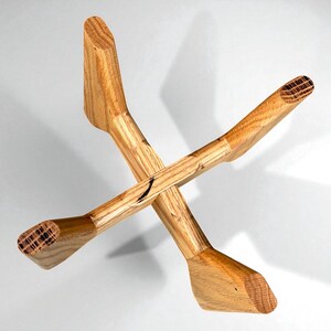 May include: A handcrafted wooden object with an X-shaped design. The natural wood has a light brown color with visible grain patterns. The ends are wider and angled, creating a unique aesthetic. The object appears to be a decorative piece.