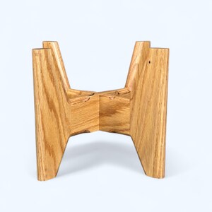 May include: A wooden plant stand with a natural oak finish. The stand has a unique, geometric design with four angled legs that intersect at the center. The stand is designed to hold a potted plant.