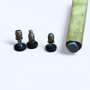 May include: Three metal screws with black bases are arranged on a white surface. A wooden furniture leg with a dark gray end cap is also visible. The screws have silver and gold tones.
