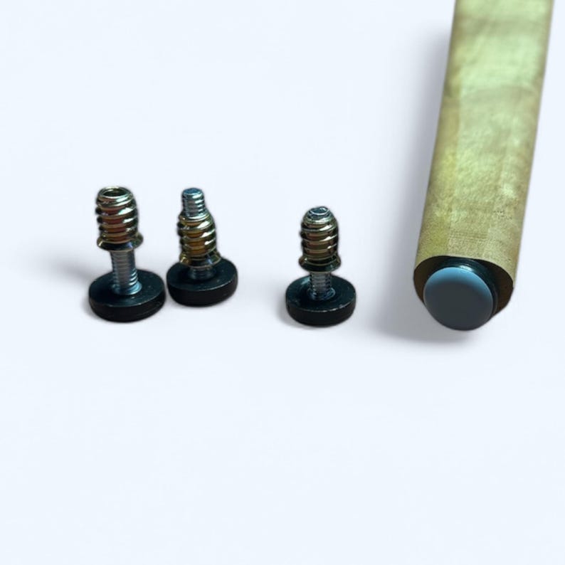 May include: Three metal screws with black bases are arranged on a white surface. A wooden furniture leg with a dark gray end cap is also visible. The screws have silver and gold tones.