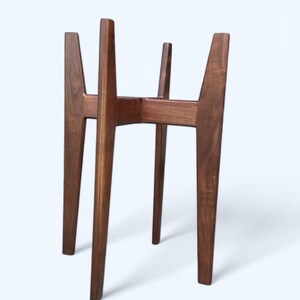 May include: A mid-century modern style plant stand made of dark brown wood. The stand has four angled legs and a central support structure, designed to hold a potted plant. The wood grain is visible.