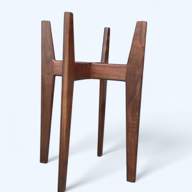 May include: A mid-century modern style plant stand made of dark brown wood. The stand has four angled legs and a central support structure, designed to hold a potted plant. The wood grain is visible.