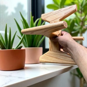 May include: A wooden plant stand with a unique, geometric design. The stand is made of light-colored wood and features multiple arms extending outwards. The stand is being held in front of a window with potted plants.