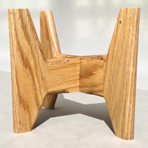 May include: A light brown wooden plant stand with a geometric design. The stand has four angled legs that meet in the center, creating a sturdy base. The wood grain is visible, adding texture to the piece. The stand is approximately 12 inches tall.