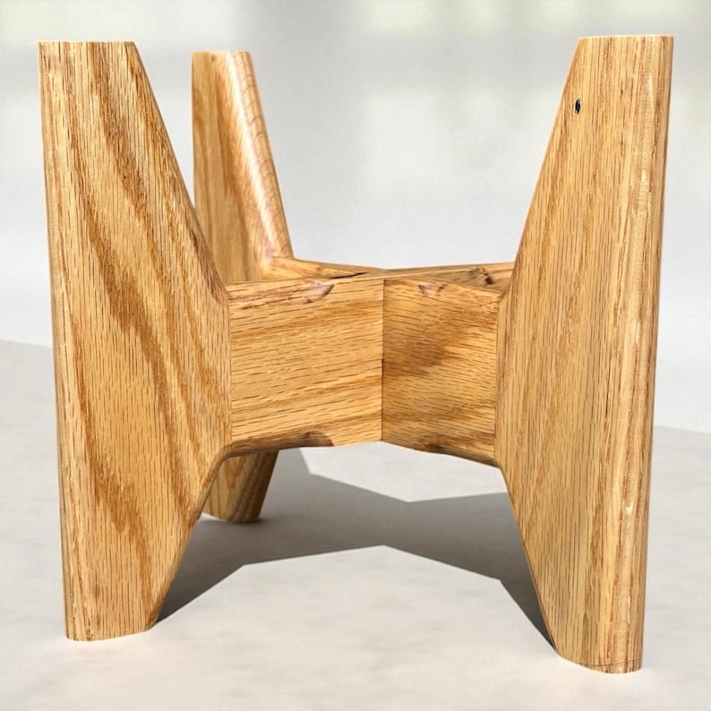 May include: A light brown wooden plant stand with a geometric design. The stand has four angled legs that meet in the center, creating a sturdy base. The wood grain is visible, adding texture to the piece. The stand is approximately 12 inches tall.