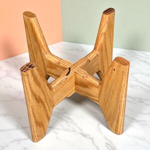 May include: A wooden plant stand with a natural finish. The stand has four angled legs that intersect in the center, creating a stable base. The wood grain is visible, adding to the stand's rustic charm. The stand is designed to hold a plant pot.