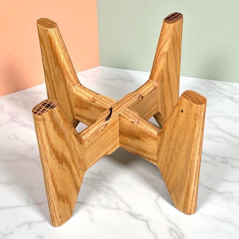 May include: A wooden plant stand with a natural finish. The stand has four angled legs that intersect in the center, creating a stable base. The wood grain is visible, adding to the stand's rustic charm. The stand is designed to hold a plant pot.