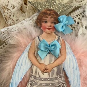 May include: A paper doll angel with pink and blue feathers, a silver star crown, and a blue bow. The doll is wearing a white dress with a blue bow and has a sweet expression.