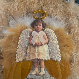 May include: A decorative angel wreath featuring a vintage-style image of a child angel with glittery wings and a gold halo. The angel is surrounded by tan feathers and a wreath base, with silver ornaments.