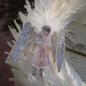 Puede incluir: Un adorno de &aacute;ngel de plumas blancas con alas cubiertas de purpurina y una mu&ntilde;eca de papel de estilo vintage de una ni&ntilde;a con un vestido blanco y sosteniendo un p&aacute;jaro.
