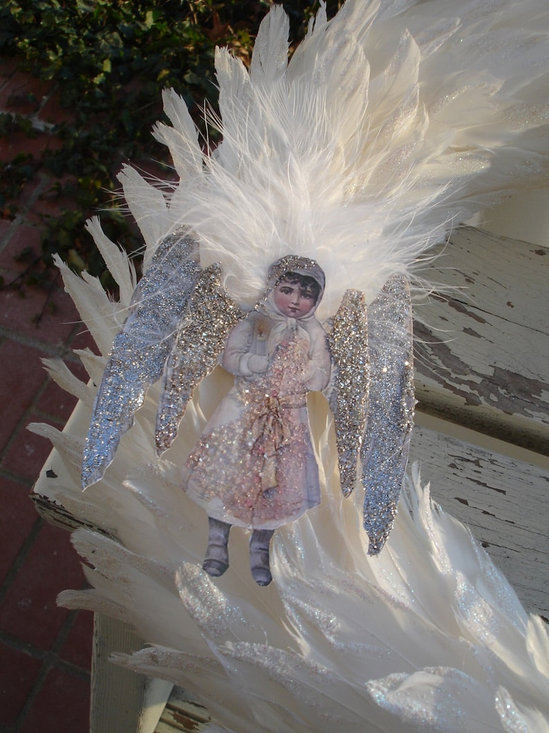 Puede incluir: Un adorno de &aacute;ngel de plumas blancas con alas cubiertas de purpurina y una mu&ntilde;eca de papel de estilo vintage de una ni&ntilde;a con un vestido blanco y sosteniendo un p&aacute;jaro.