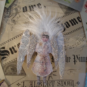 Puede incluir: Una figurita de &aacute;ngel de estilo vintage con alas de purpurina plateada y un halo de plumas blancas. El &aacute;ngel lleva un vestido blanco con estampado floral rosa y sostiene un peque&ntilde;o objeto blanco en sus manos. La figurita est&aacute; sentada sobre un trozo de papel con el texto "J. Albert Snow, Author of "Old Oaken Bucket;" Variations "Blossom Waltzes;" Elu" impreso en &eacute;l.