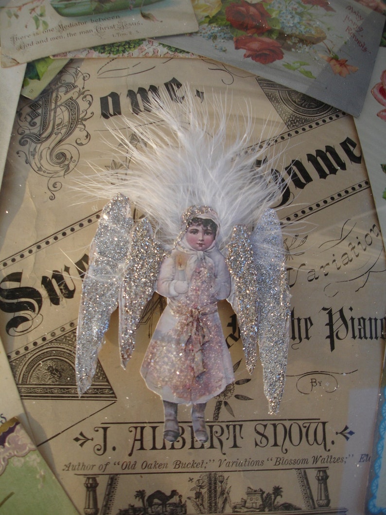 Puede incluir: Una figurita de &aacute;ngel de estilo vintage con alas de purpurina plateada y un halo de plumas blancas. El &aacute;ngel lleva un vestido blanco con estampado floral rosa y sostiene un peque&ntilde;o objeto blanco en sus manos. La figurita est&aacute; sentada sobre un trozo de papel con el texto "J. Albert Snow, Author of "Old Oaken Bucket;" Variations "Blossom Waltzes;" Elu" impreso en &eacute;l.