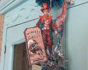 May include: A whimsical circus-themed ornament featuring a figure in a red and orange suit, a giraffe, and a sign that reads "Wonders of the Circus." The ornament includes a blue glitter ball base and a small blue umbrella. The sign has the number N0039883.