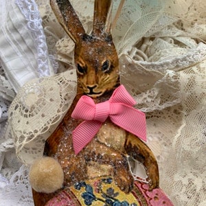 May include: A vintage Easter bunny ornament made of paper with a pink bow and holding a pink and yellow Easter egg with floral designs.