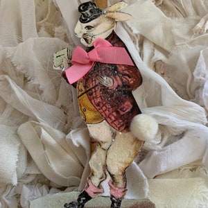 May include: A vintage paper mache ornament of a rabbit wearing a top hat, a pink bow tie, and a red and yellow jacket. The ornament is decorated with glitter and has a white cotton tail.