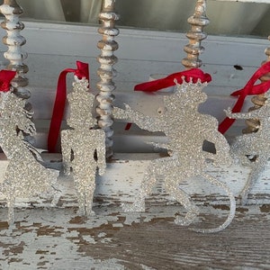 May include: Four silver glitter ornaments shaped like a ballerina, a nutcracker, a prince, and a mouse. The ornaments are hanging from red ribbon.