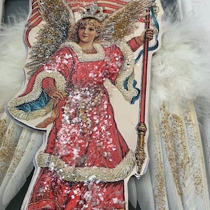 May include: A vintage-style angel figurine holding an American flag. The angel is wearing a red dress with white fur trim and a gold crown. The wings are made of white feathers with gold glitter accents. The flag is red, white, and blue with gold stars. The figurine is decorated with white glitter.