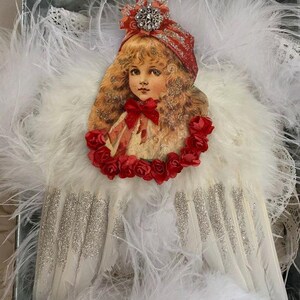 May include: A vintage-inspired angel ornament with white feathery wings and a red and white paper doll head with a red bow. The doll has blonde hair and blue eyes. The ornament is decorated with red roses.