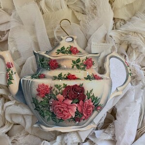 May include: A decorative teapot ornament with a cream-colored base and a floral design featuring red and pink roses. The teapot has a gold-colored hook for hanging and is embellished with glitter.