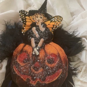 May include: A witch fairy figurine with black wings and a black hat sits on a glittery orange pumpkin. The fairy has blonde hair and is wearing a black dress with white socks and black shoes.