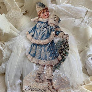 May include: Vintage-style Christmas ornament featuring a child in a blue winter coat and hat, holding a small fir tree. The ornament has a glittery finish and the word "Greetings" at the bottom. The background includes white feathers and fabric.