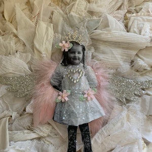 May include: A handmade angel ornament with pink feathers, silver glitter wings, and a black and white photo of a young girl wearing a crown and a sparkly dress.