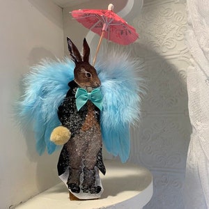 May include: A brown rabbit wearing a black suit with a blue bow tie and blue feather wings, holding a pink umbrella.