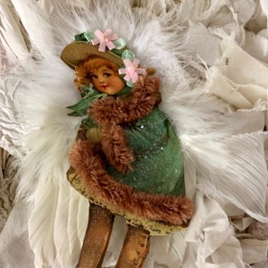 May include: A vintage Christmas ornament featuring a girl wearing a green dress, brown fur trim, and a hat with pink flowers. The ornament has white feathers and is surrounded by white fabric.
