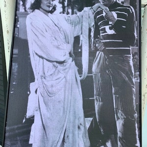 May include: A black and white photograph of a woman in a long white robe and a man in a striped shirt and trousers. The woman is standing with her hand on the man's shoulder, and the man is looking down at her.