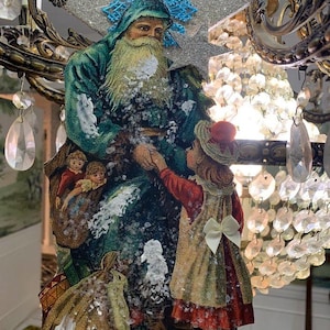 May include: A vintage Christmas ornament featuring Santa Claus in a green robe, holding hands with a girl in a red dress. The ornament has a glittery star and blue snowflake accents. A sack of toys is at Santa's feet.