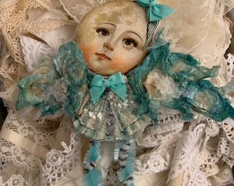 May include: A handmade moon-shaped ornament with a painted face, wearing a blue and white dress with a blue bow. The ornament is surrounded by white lace.