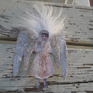 Puede incluir: Un adorno de &aacute;ngel de plumas blancas con alas de purpurina plateada y una mu&ntilde;eca de papel con un vestido blanco y un lazo rojo. La mu&ntilde;eca sostiene un objeto blanco en sus manos.