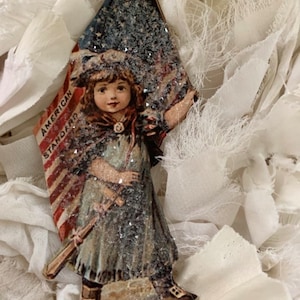 May include: A vintage-style Christmas ornament featuring a girl holding an American flag. The ornament is made of paper and has a glittery finish. The text "AMERICAN STANDARD" is printed on the flag.