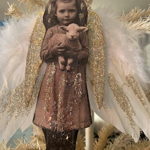 May include: A vintage-style Christmas ornament featuring a sepia-toned image of a girl holding a lamb. The ornament has feathered white wings with gold glitter accents. The girl wears a brown dress and a crown. The ornament is designed to hang on a Christmas tree.