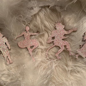 May include: Five pink glitter ornaments depicting characters from the Nutcracker ballet. The ornaments include a ballerina, a nutcracker soldier, a mouse king, and two other characters.