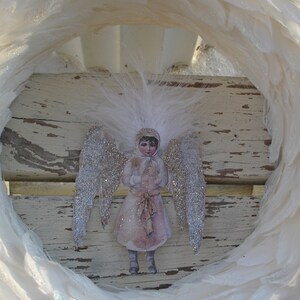Puede incluir: Un adorno de &aacute;ngel de plumas blancas con alas de brillantina y un vestido blanco. El &aacute;ngel sostiene un peque&ntilde;o objeto blanco en sus manos. El adorno est&aacute; colgado de una corona blanca.