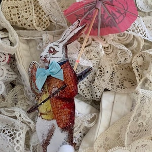 Puede incluir: Un conejo blanco con una pajarita azul y una chaqueta roja sostiene un palo con una sombrilla de papel rosa. El conejo está rodeado de tela de encaje blanca.