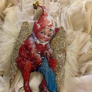 May include: A vintage-style Christmas bauble featuring a red and white elf with gold glitter wings. The elf is holding a blue stocking and has a bell on its hat.