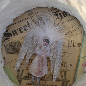 Puede incluir: Un adorno navide&ntilde;o vintage con un &aacute;ngel de purpurina plateada con alas blancas y un halo de plumas blancas. El &aacute;ngel lleva un vestido blanco con una cinta roja y sostiene una flor blanca. El adorno est&aacute; colocado sobre un fondo de una p&aacute;gina de m&uacute;sica vintage con el texto "Sweet Aviation" y "By J.W.  from Waltzes."