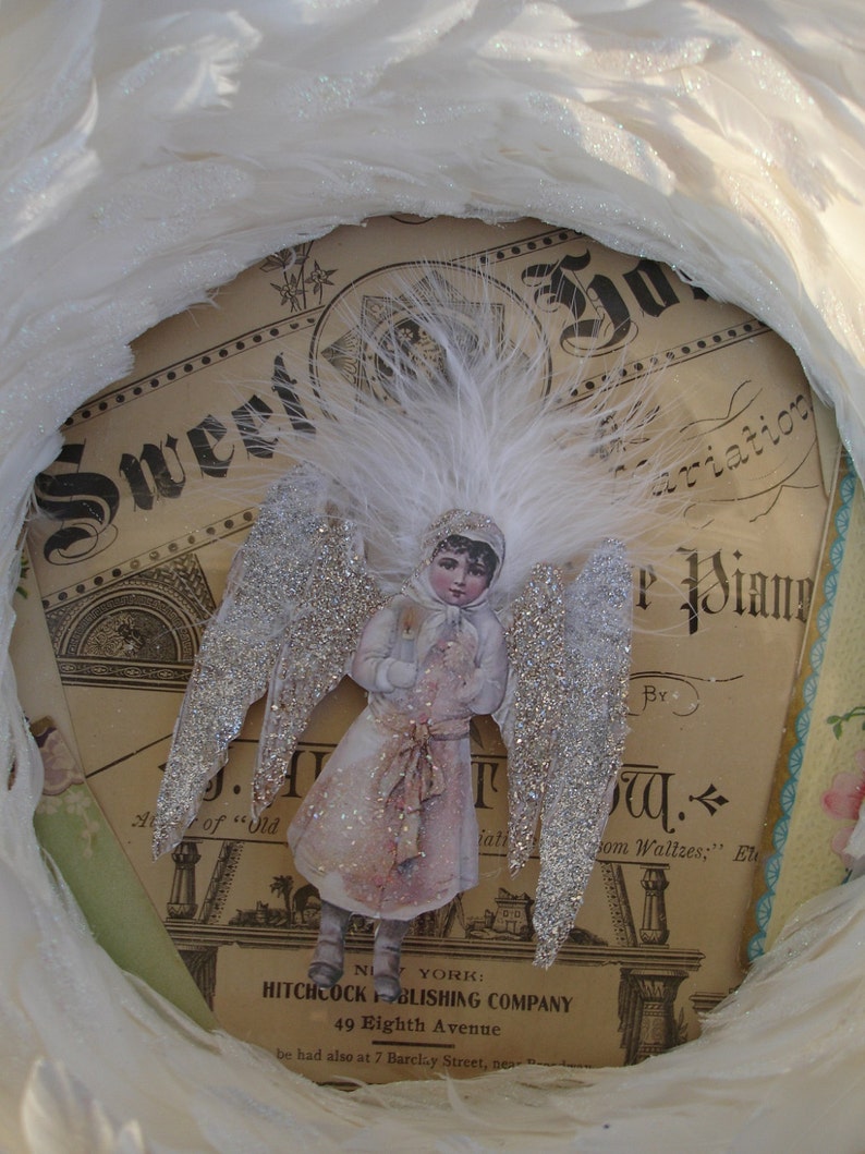 Puede incluir: Un adorno navide&ntilde;o vintage con un &aacute;ngel de purpurina plateada con alas blancas y un halo de plumas blancas. El &aacute;ngel lleva un vestido blanco con una cinta roja y sostiene una flor blanca. El adorno est&aacute; colocado sobre un fondo de una p&aacute;gina de m&uacute;sica vintage con el texto "Sweet Aviation" y "By J.W.  from Waltzes."