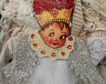 May include: A vintage-style Christmas ornament featuring a heart-shaped head with a crown, a child's face, and silver glitter wings. The ornament is decorated with red and white hearts.