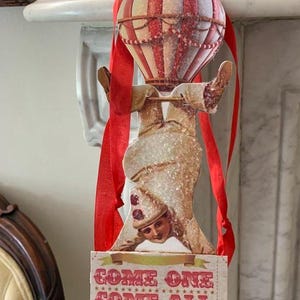 Puede incluir: Adorno vintage con temática de circo con un globo aerostático a rayas rojas y blancas y un artista. El adorno tiene una cinta roja para colgar y un letrero que dice "COME ONE COME ALL".
