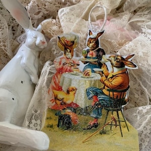 May include: A vintage-style Easter ornament featuring four bunnies dressed in clothing, sitting at a table with a chick and a hat. The ornament is made of wood and has a glittery finish.