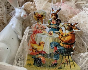 May include: A vintage-style Easter ornament featuring four bunnies dressed in clothing, sitting at a table with a chick and a hat. The ornament is made of wood and has a glittery finish.