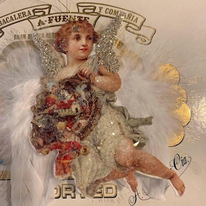 May include: A vintage-style angel ornament with white feather wings and glitter accents. The angel figure is holding a decorative basket. The ornament has a gold string for hanging and is set against a background with text.