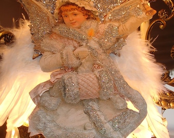 May include: A white angel figurine with silver glitter accents, holding a candle and wearing a white dress with a pink sash. The angel is perched on a crystal chandelier.