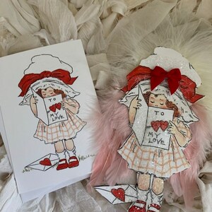 May include: A vintage Valentine's Day card featuring a girl in a red bow and white dress holding a card that says "To My Love". The card is surrounded by white feathers.