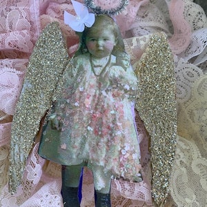 May include: A vintage-style angel ornament with glitter wings, a floral dress, and a halo. The angel is made of paper and has a delicate, whimsical look.